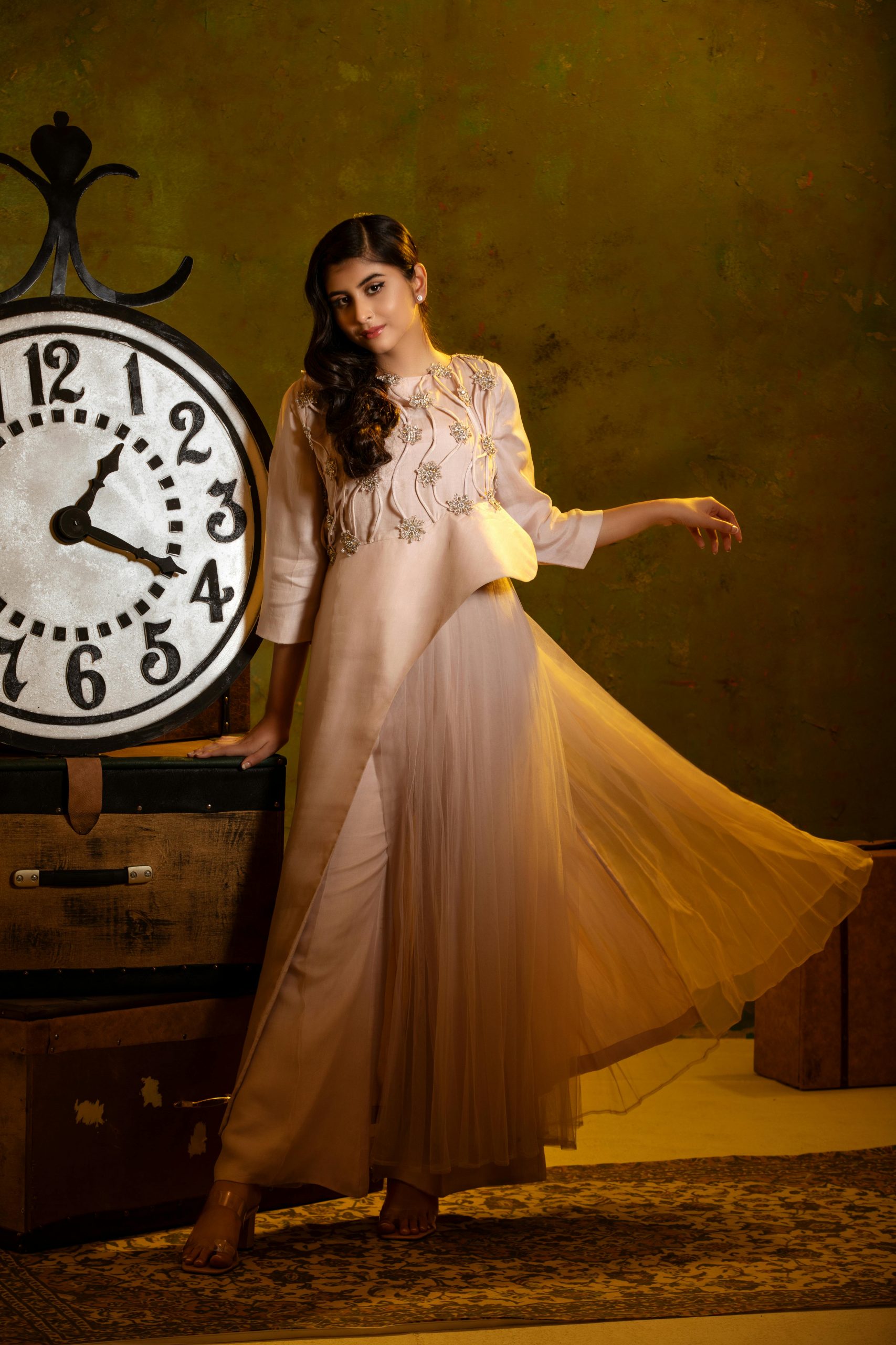 A woman in a flowing dress poses elegantly beside a vintage clock indoors.